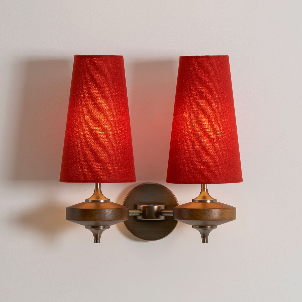 Red shades lit warm from inside on a Clérachic twin sconce, with walnut discs and a bronze link arm, on a soft grey wall.