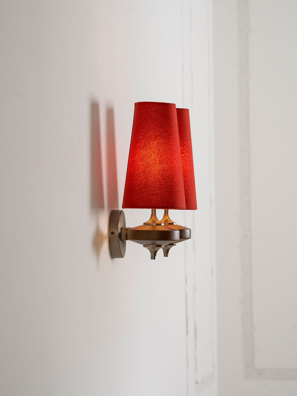 Side-on view of a Clérachic wall sconce with two red shades beside a white panelled door frame.