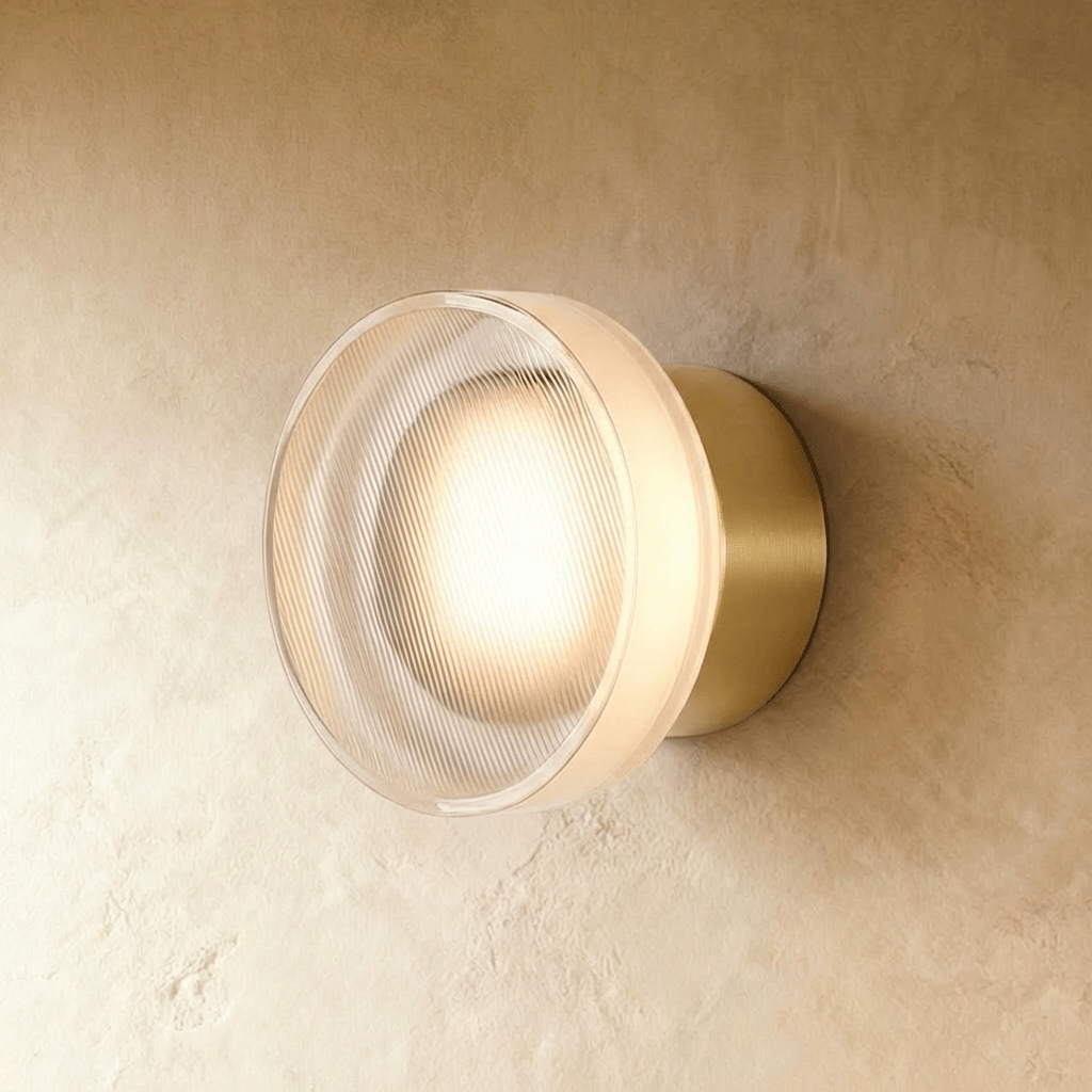 Clérachic round wall light with a ribbed clear glass shade glowing warm, on a brushed gold wall plate against limewash plaster.