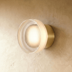 Clérachic round wall light with a ribbed clear glass shade glowing warm, on a brushed gold wall plate against limewash plaster.