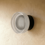 Clérachic round wall light switched off, showing its finely ribbed glass shade on a matt black wall plate and limewash plaster.