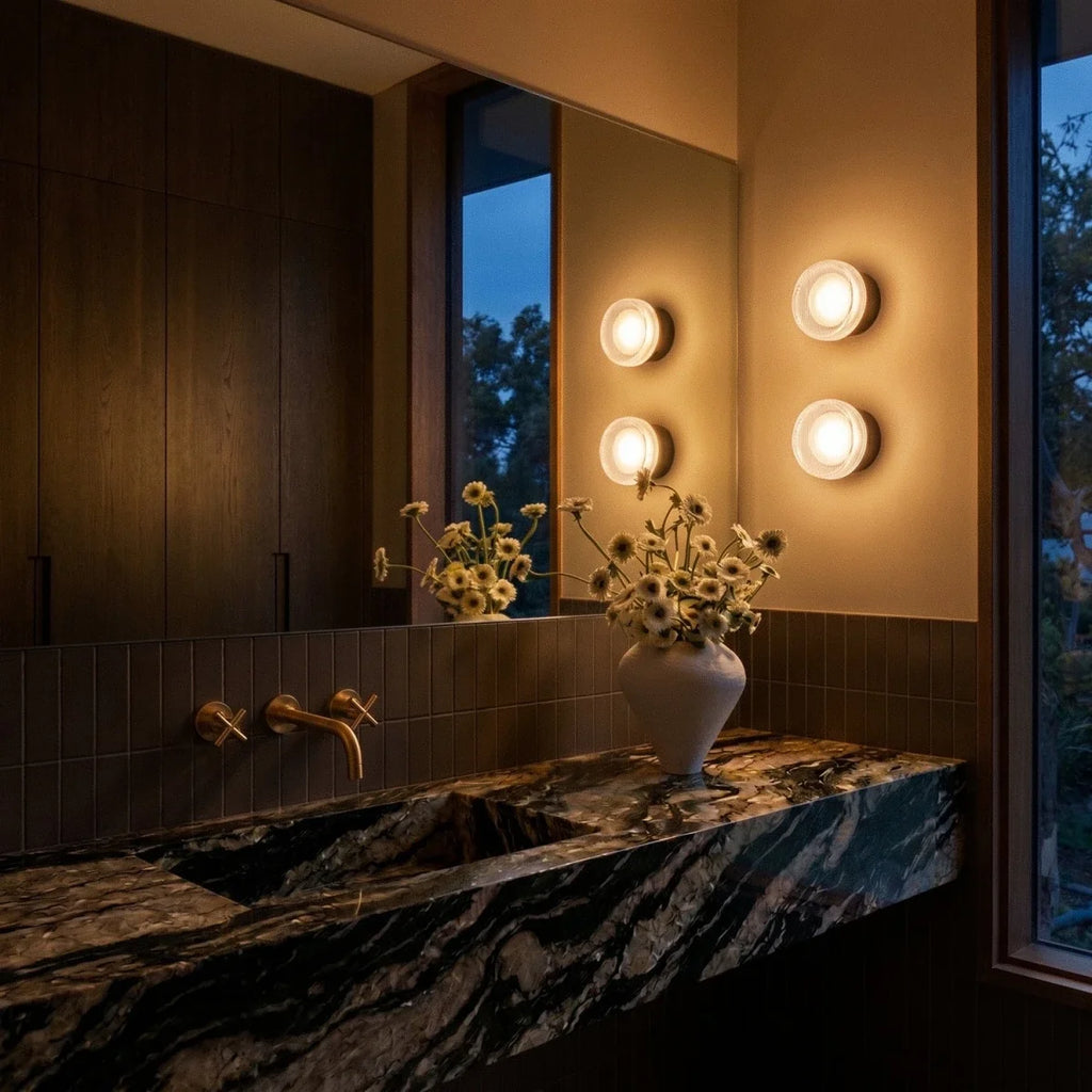 Clérachic round ribbed-glass wall lights lit at dusk beside a bathroom mirror, with a vase of daisies on the marble basin.
