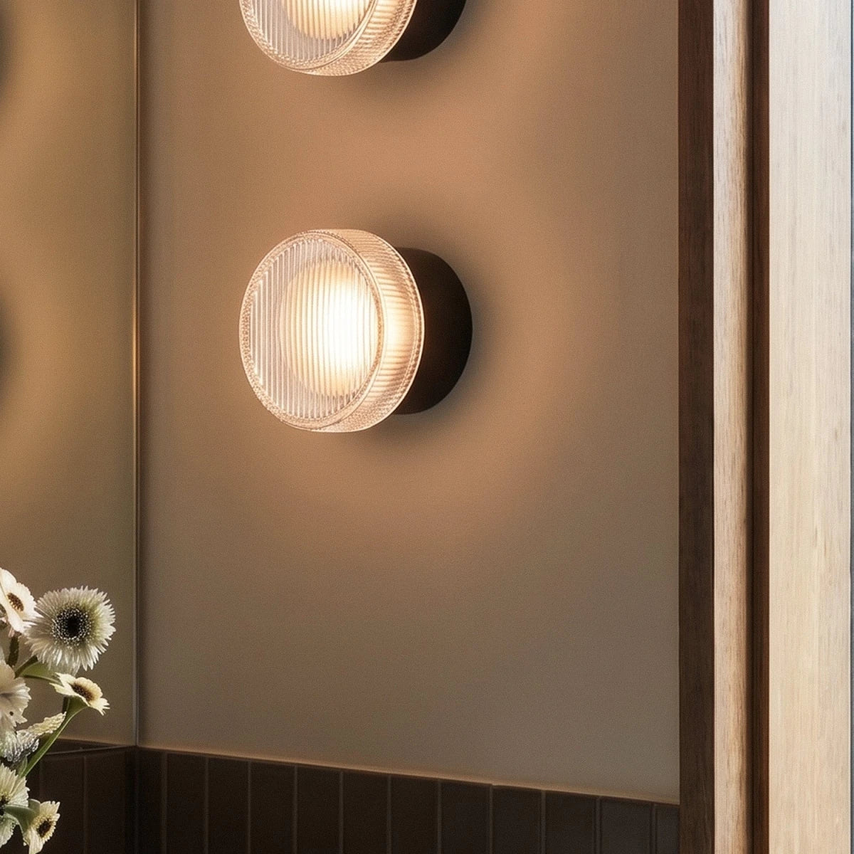 Close view of a Clérachic round wall light with a ribbed glass shade and black wall plate, glowing warm on a plain wall.
