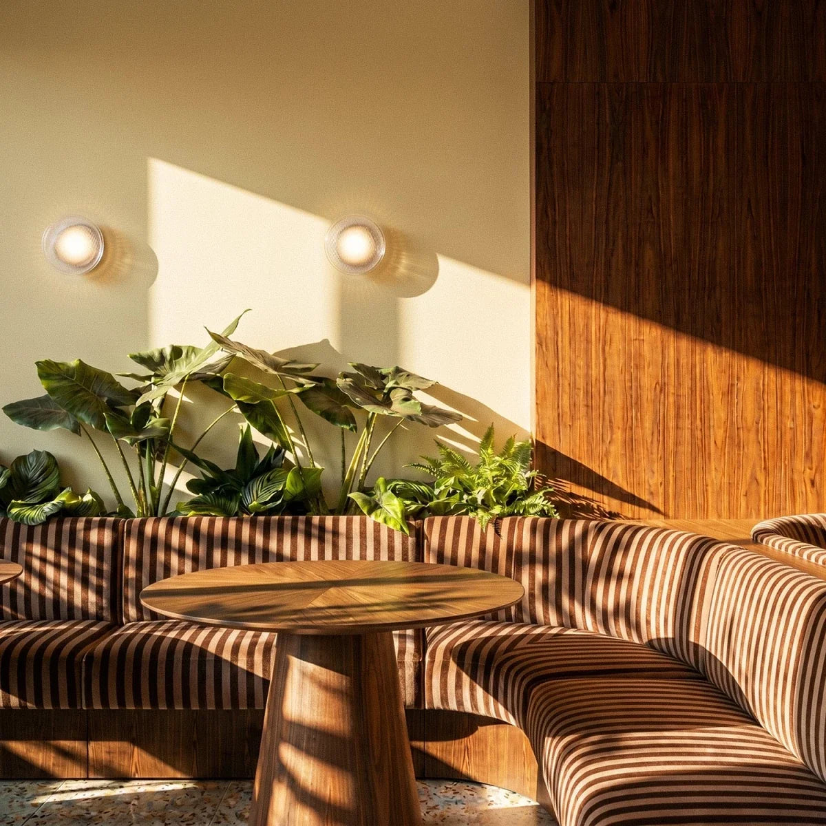 Two Clérachic round wall lights glowing in afternoon sun above leafy plants and a striped banquette with a walnut table.