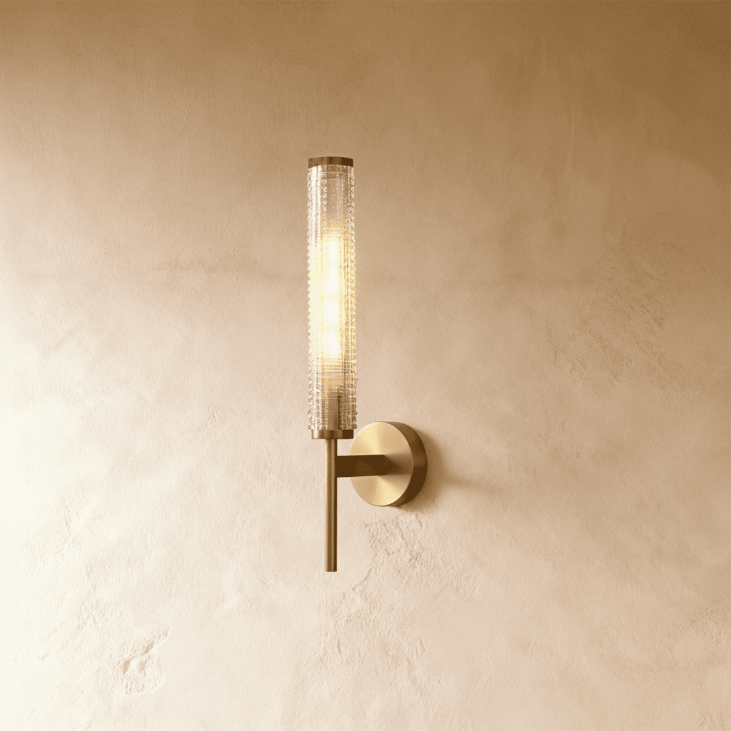Clérachic brass wall sconce with a slim round-based plate and a textured clear glass cylinder, lit warm on a limewashed wall.