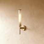 Clérachic brass wall sconce with a slim round-based plate and a textured clear glass cylinder, lit warm on a limewashed wall.