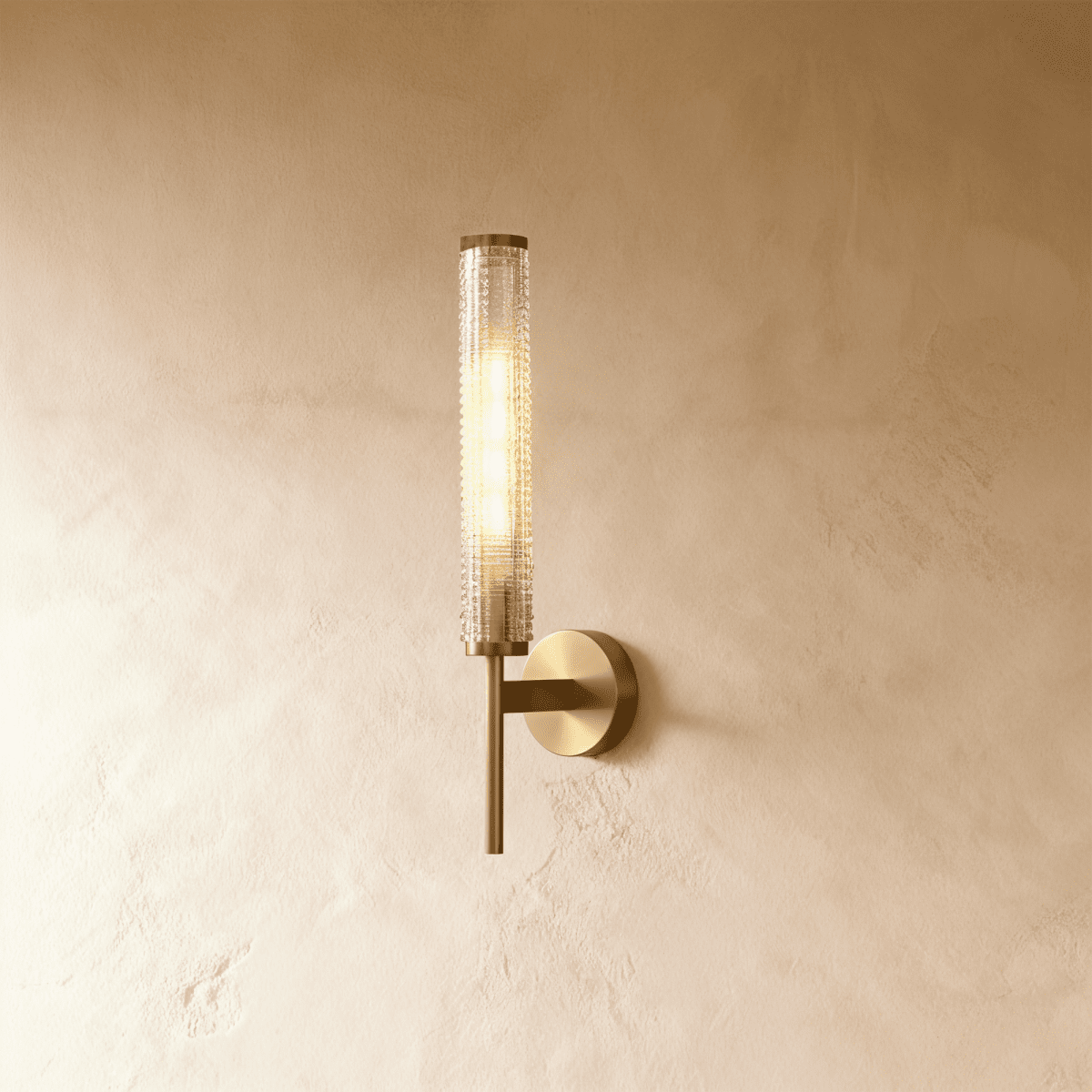 Clérachic brass wall sconce with a slim round-based plate and a textured clear glass cylinder, lit warm on a limewashed wall.