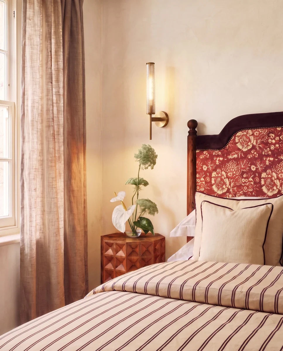 Clérachic brass wall sconce glowing beside a bed with a floral headboard and striped linen, against a soft plaster wall.