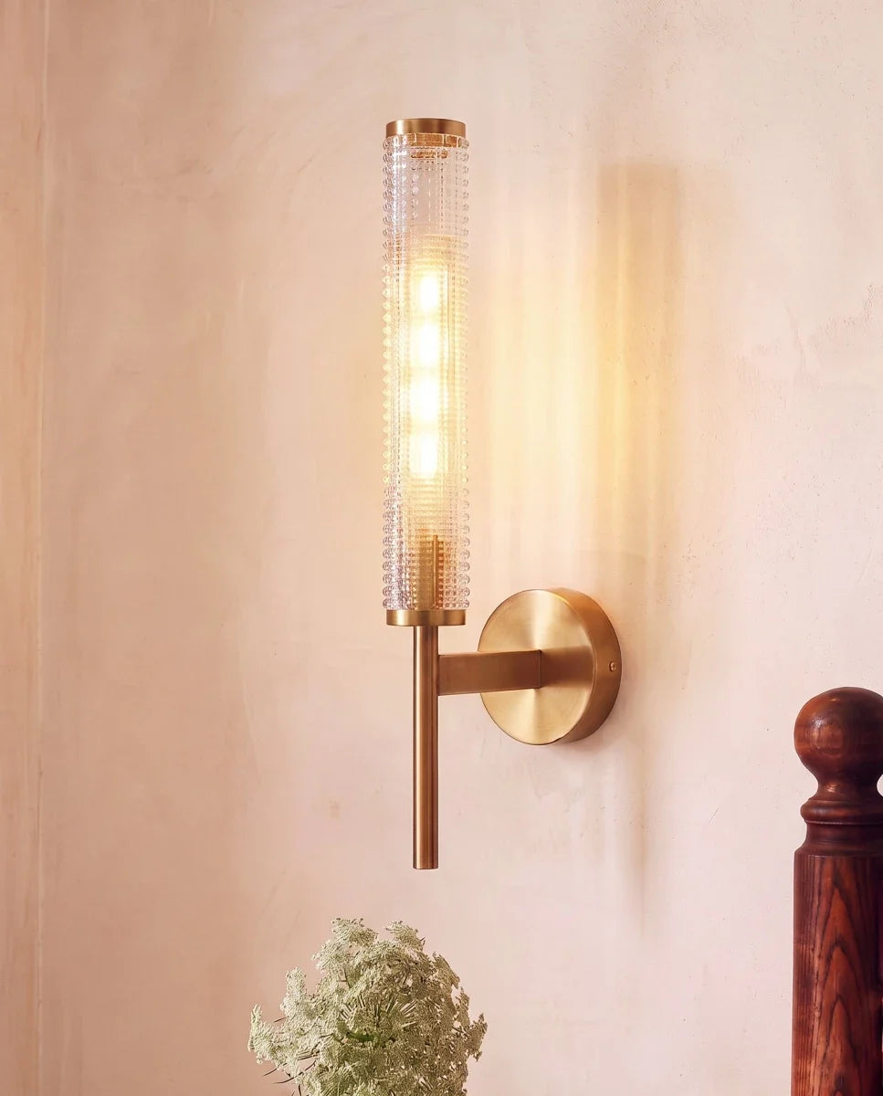 Clérachic lit brass wall sconce with a beaded glass cylinder, mounted on a blush plaster wall above a spray of white flowers.