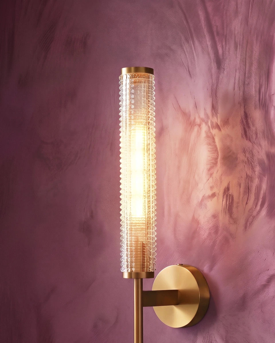 Clérachic brass wall sconce lit warm against a deep plum plaster wall, its beaded glass cylinder glowing from within.