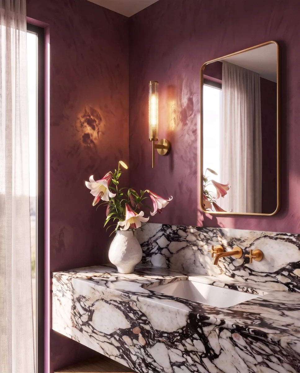 Clérachic brass wall sconce lit beside a gilt mirror in a plum-walled bathroom with a marble basin and brass taps.