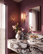Clérachic brass wall sconce lit beside a gilt mirror in a plum-walled bathroom with a marble basin and brass taps.