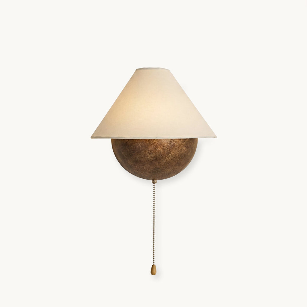 Clérachic wall light with a tapered fabric shade over a rounded iron half-sphere and a brass pull chain, on a plain white backdrop.