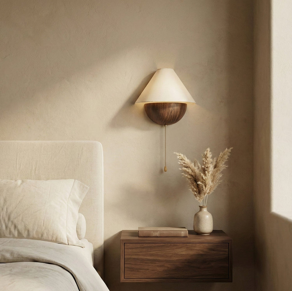 Lit Clérachic wall light above a floating walnut nightstand, beside a bed and a vase of dried pampas grass.