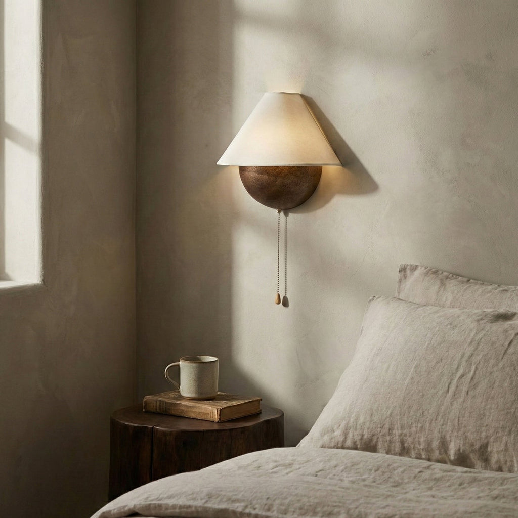 Clérachic wall light mounted beside a bed in soft window light, with a mug and a book on a log-style bedside table.