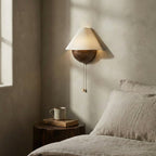 Clérachic wall light mounted beside a bed in soft window light, with a mug and a book on a log-style bedside table.