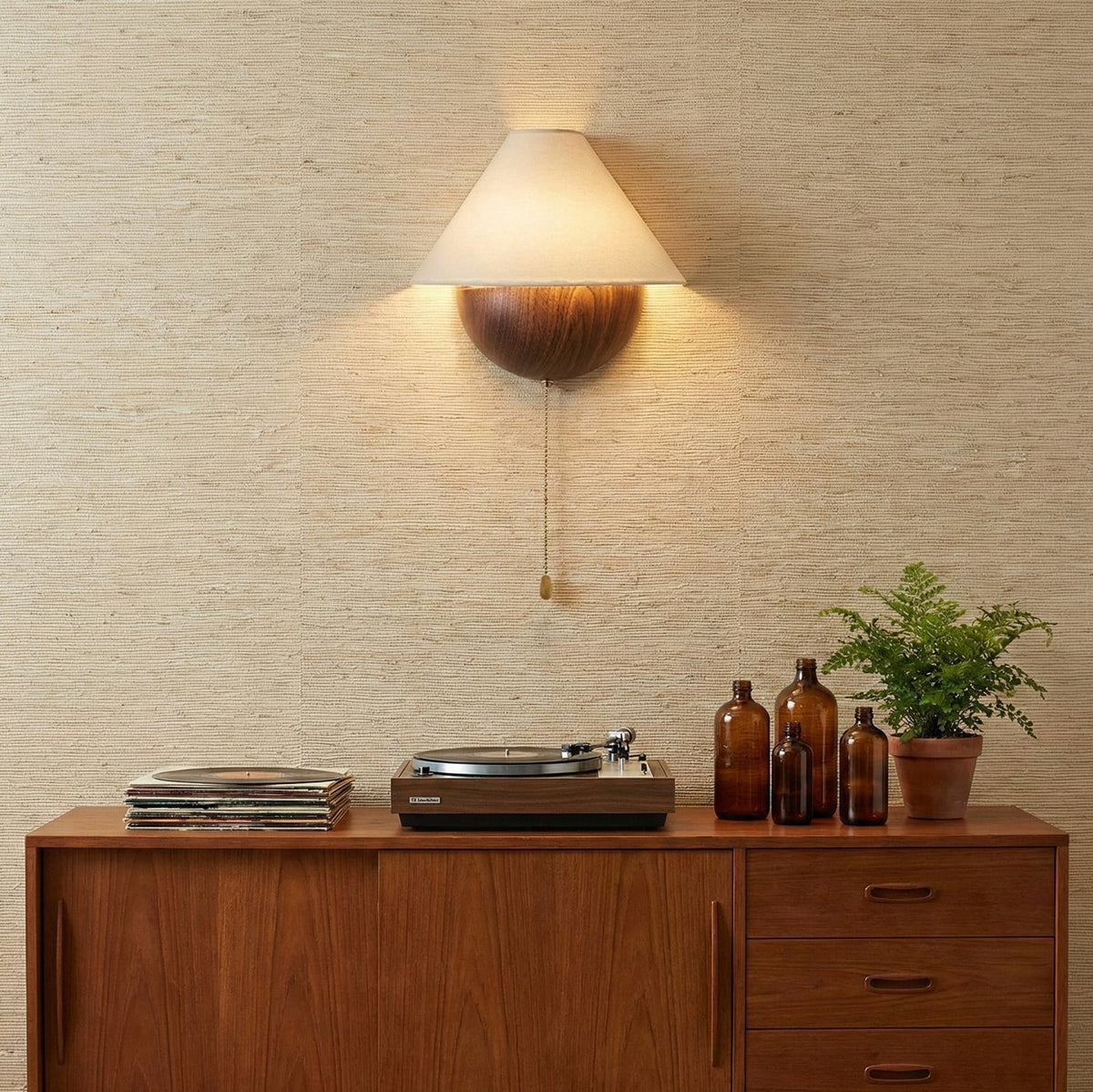 Clérachic wall light glowing over a teak sideboard that holds a record player, amber bottles and a small fern.