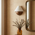 Clérachic wall light on a grass-cloth wall beside a gilt-edged mirror, with eucalyptus stems in an amber glass vase.