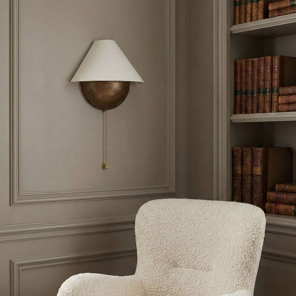 Clérachic wall light on a grey panelled wall, beside a cream bouclé armchair and a shelf of leather-bound books.