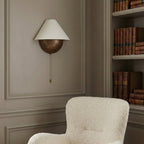 Clérachic wall light on a grey panelled wall, beside a cream bouclé armchair and a shelf of leather-bound books.
