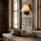 Unlit Clérachic wall light against weathered grey planks beside a window, above a table of stacked stoneware plates and bowls.