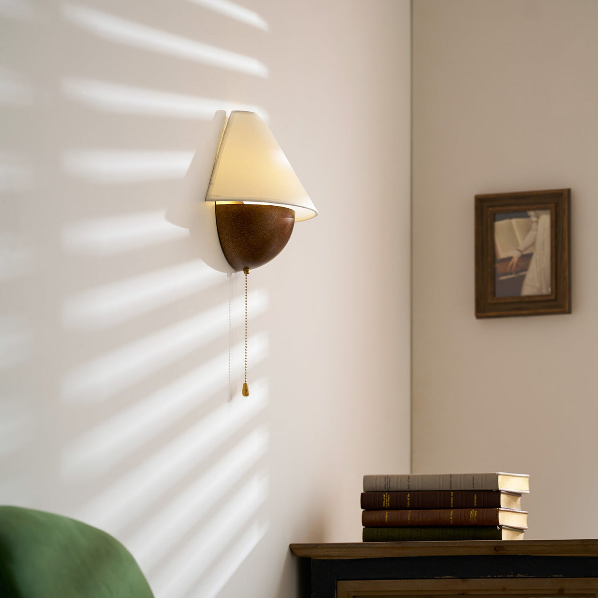 Clérachic wall light lit in striped afternoon sunlight, with a framed picture and a stack of books on a cabinet below.