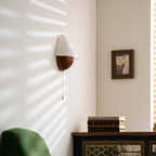 Clérachic wall light on a white wall with its pull chain hanging down, a green armchair and a small frame in view.
