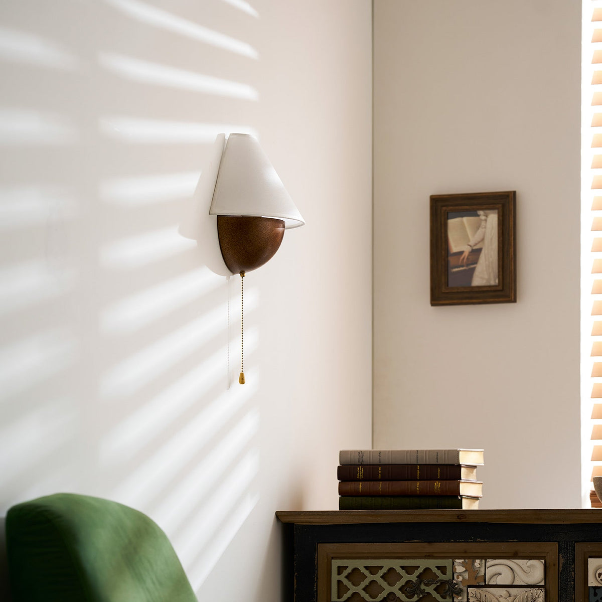 Clérachic wall light on a white wall with its pull chain hanging down, a green armchair and a small frame in view.