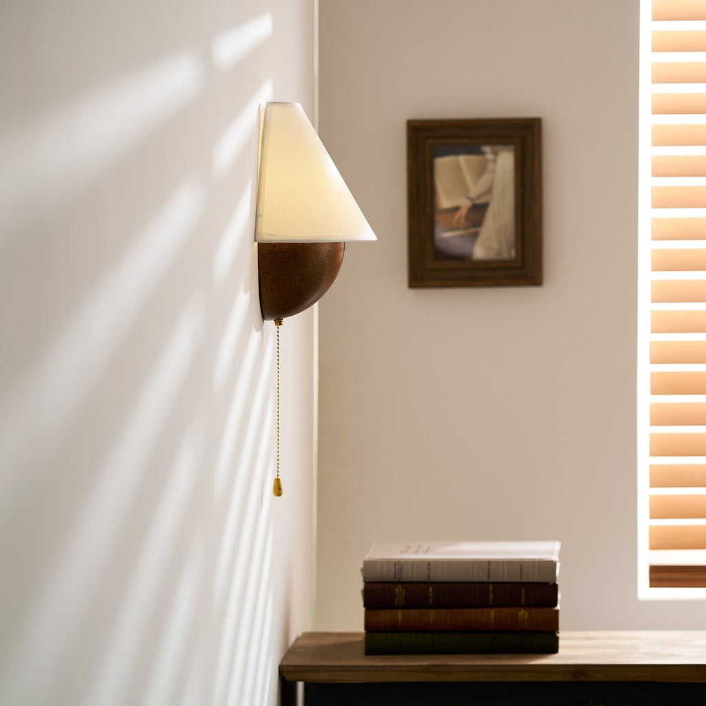 Close crop of this Clérachic wall light and its brass-tipped pull chain, with a framed picture and window blind behind.