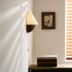 Close crop of this Clérachic wall light and its brass-tipped pull chain, with a framed picture and window blind behind.