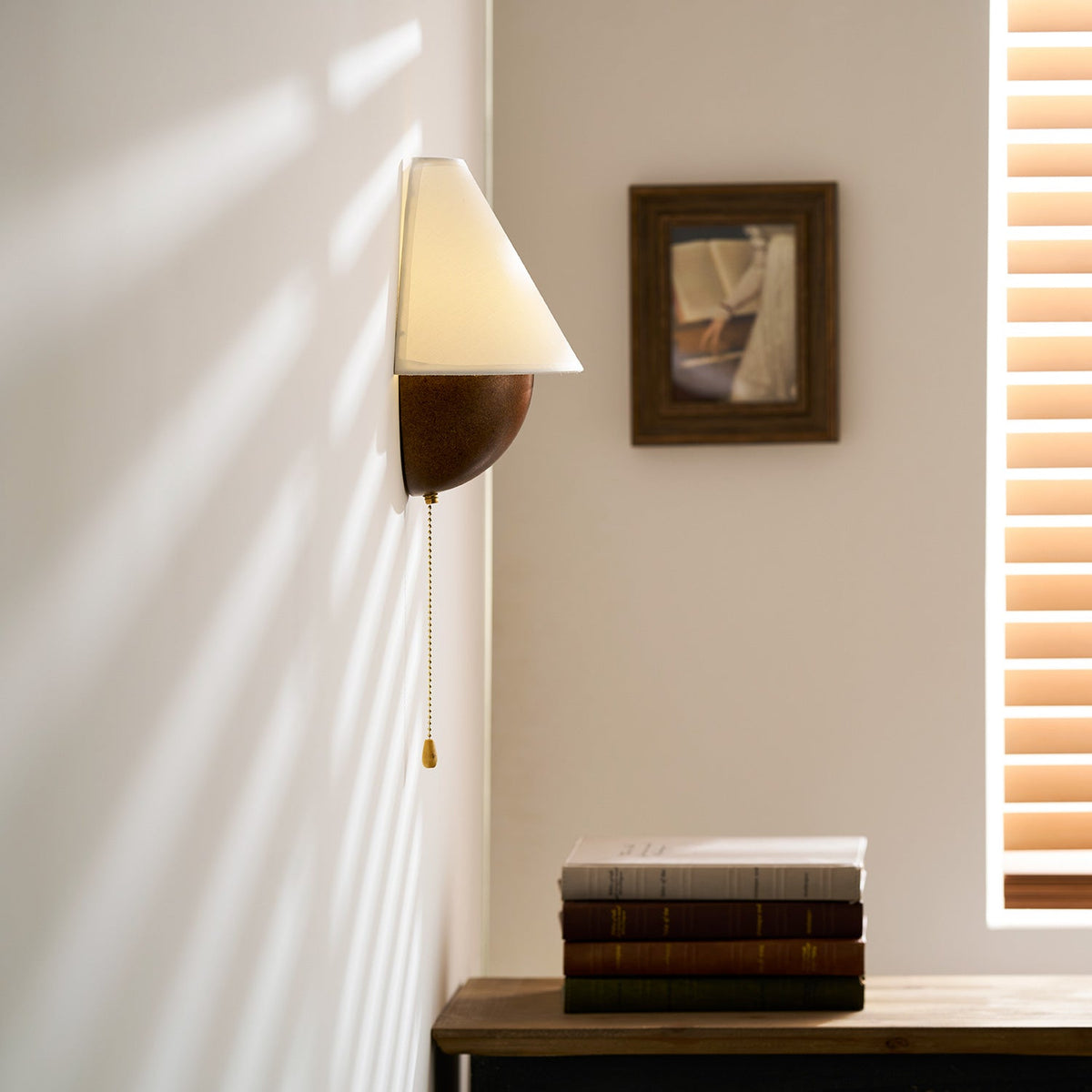 Close crop of this Clérachic wall light and its brass-tipped pull chain, with a framed picture and window blind behind.