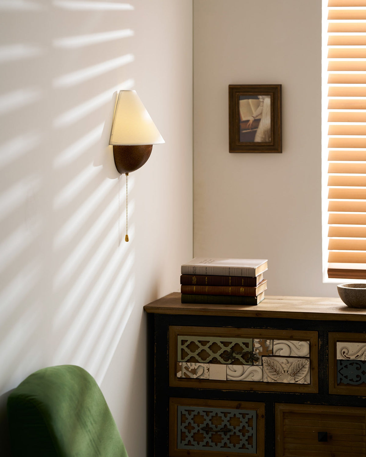 Clérachic wall light in sunlight, with a patterned wooden chest below and a green armchair at the edge of the frame.
