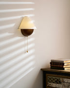 Lit Clérachic wall light in a sunbeam, with the glow reaching under the shade and a stack of books on a cabinet below.