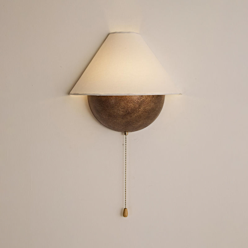 Front view of a lit Clérachic wall light on a cream wall, its brass pull chain and teardrop pull hanging below.