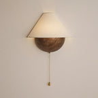 Front view of a lit Clérachic wall light on a cream wall, its brass pull chain and teardrop pull hanging below.