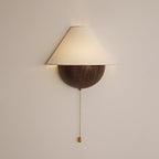 Clérachic wall light in a walnut wood-grain finish, softly lit against a pale blush wall, with its brass pull chain below.
