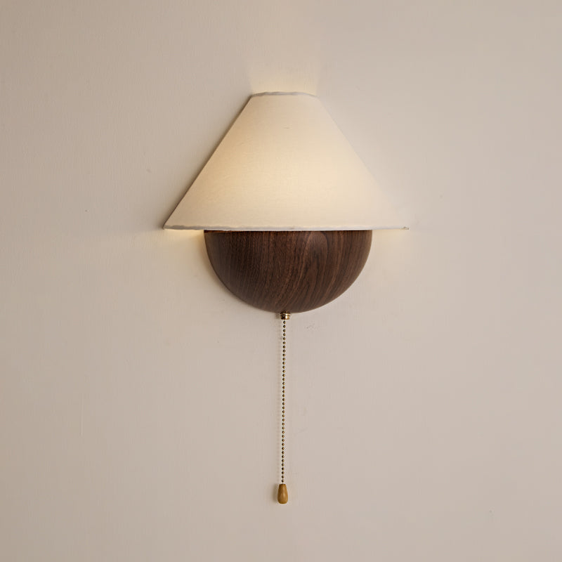 Clérachic wall light in a walnut wood-grain finish, softly lit against a pale blush wall, with its brass pull chain below.