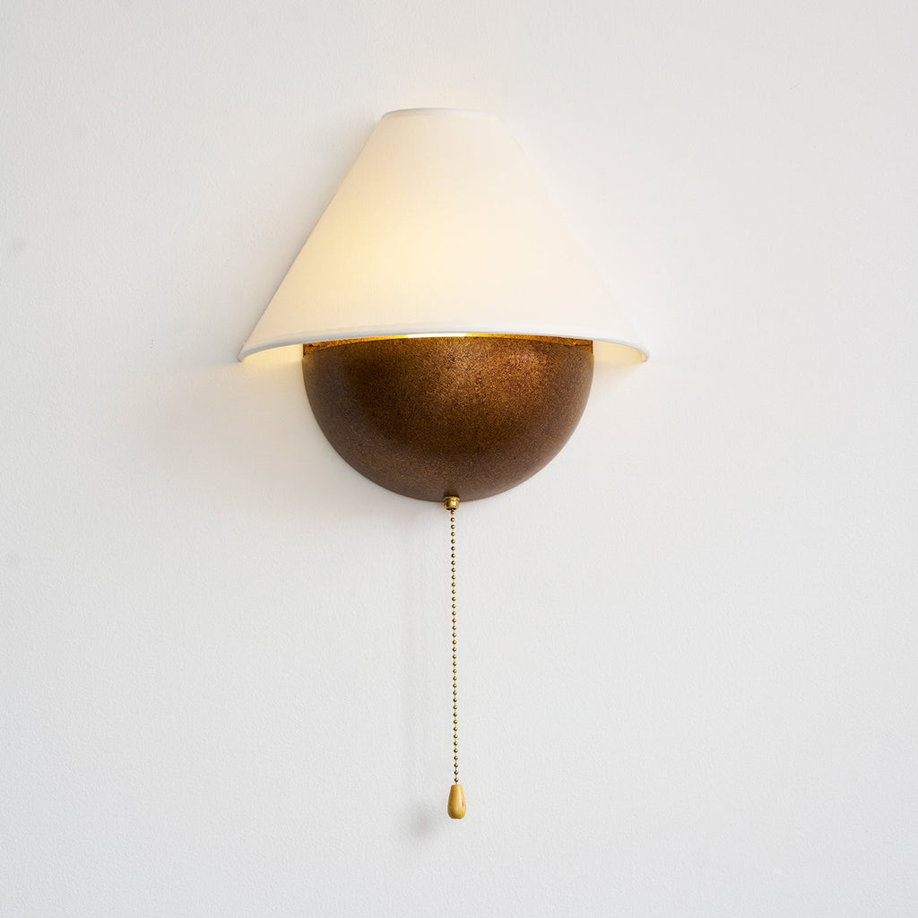 Lit Clérachic wall light in an aged bronze finish on a white wall, with a long beaded pull chain hanging down.