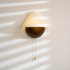 Clérachic wall light with a warm glow under the shade, in bands of afternoon sun, its beaded chain ending in a brass pull.