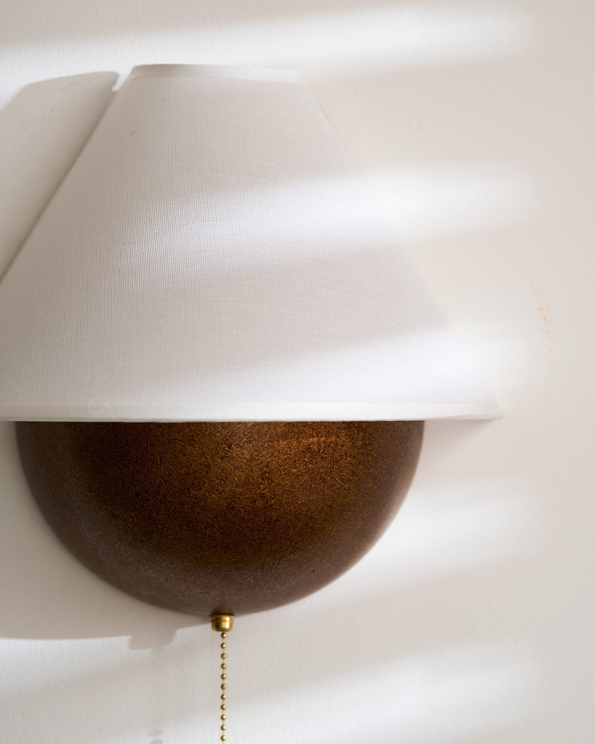 Close view of the fabric shade and textured bronze half-sphere on this Clérachic wall light, with sun stripes across it.