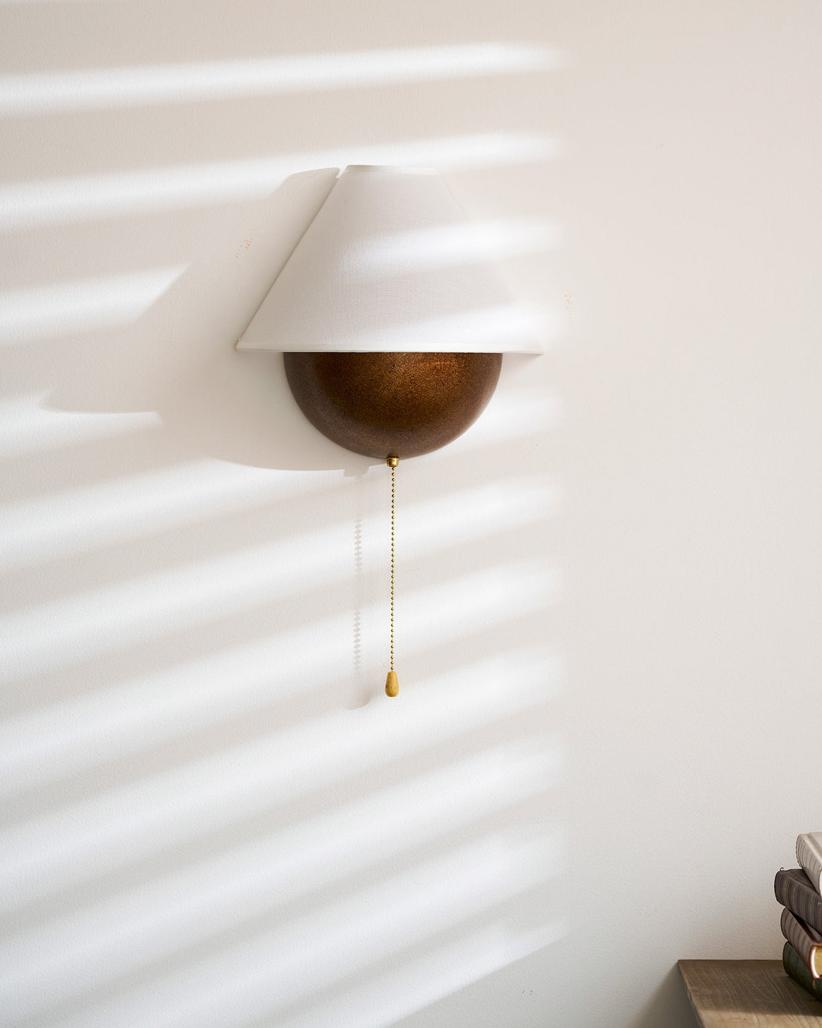 Clérachic wall light seen small on a sunlit wall, with its pull chain hanging and a stack of books at the edge.