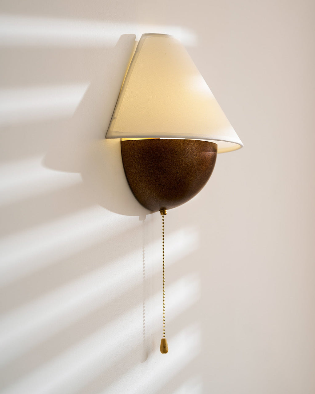 Clérachic wall light lit in sun stripes, with the shade tilted forward and a long chain hanging from the base.