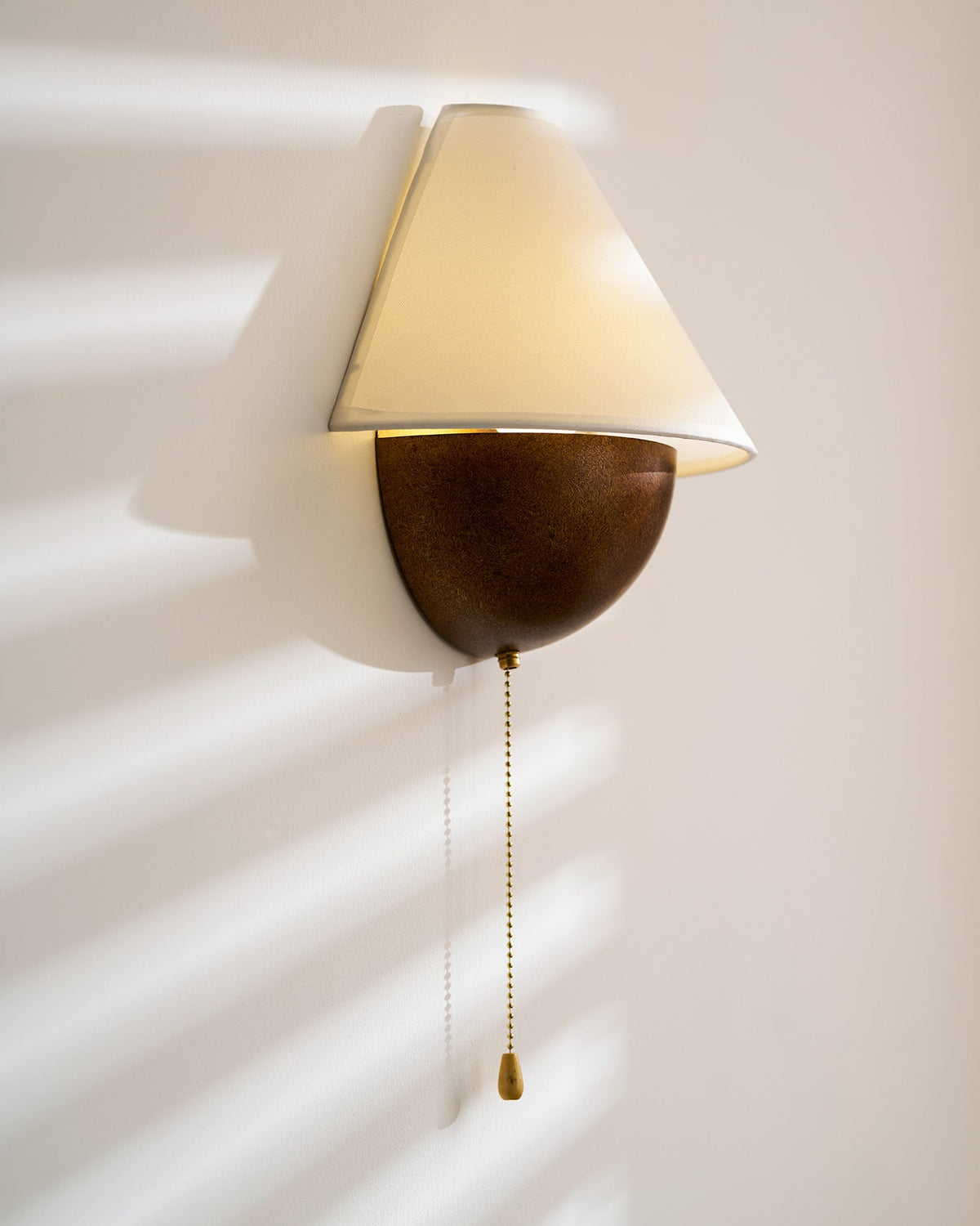 Clérachic wall light lit in sun stripes, with the shade tilted forward and a long chain hanging from the base.