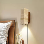 Square-sided travertine wall light from Clérachic beside a wooden bed, its downlight falling over a small vase of flowers.