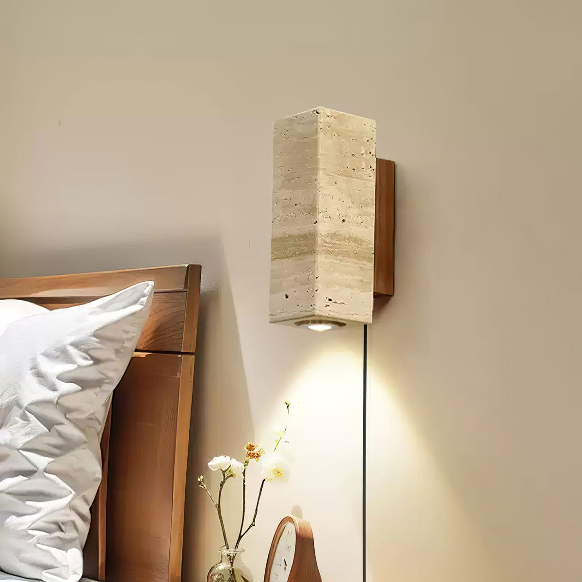 Square-sided travertine wall light from Clérachic beside a wooden bed, its downlight falling over a small vase of flowers.
