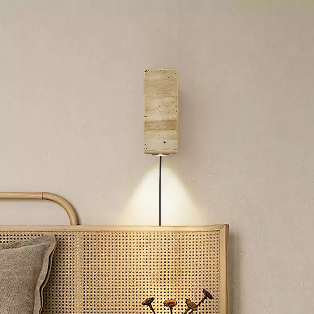 Rectangular travertine Clérachic wall light with a black cord, shining a cone of warm light down onto a rattan bedhead.