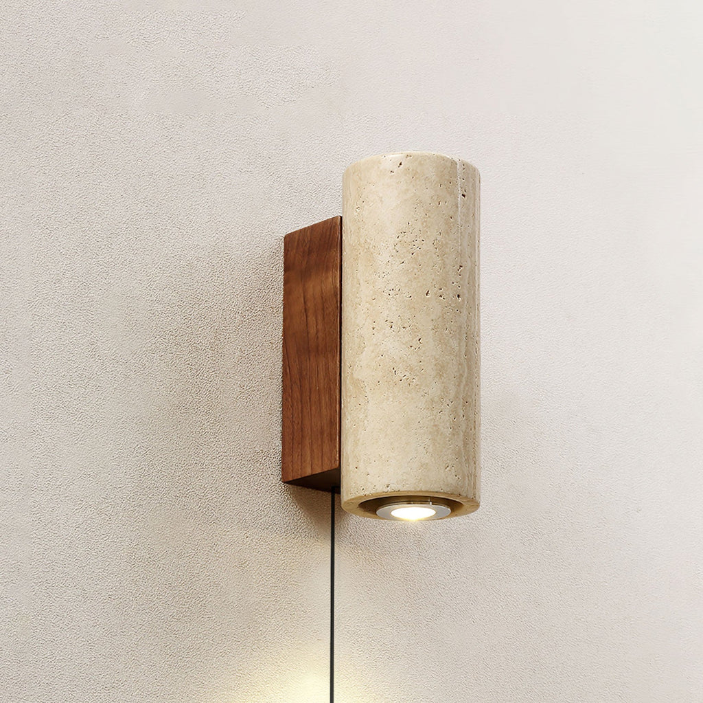 Cylindrical Clérachic wall light in pale travertine, set on a walnut-colour backplate on a plaster wall, its downlight on.