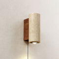 Cylindrical Clérachic wall light in pale travertine, set on a walnut-colour backplate on a plaster wall, its downlight on.