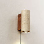 Cylindrical Clérachic wall light in pale travertine, set on a walnut-colour backplate on a plaster wall, its downlight on.