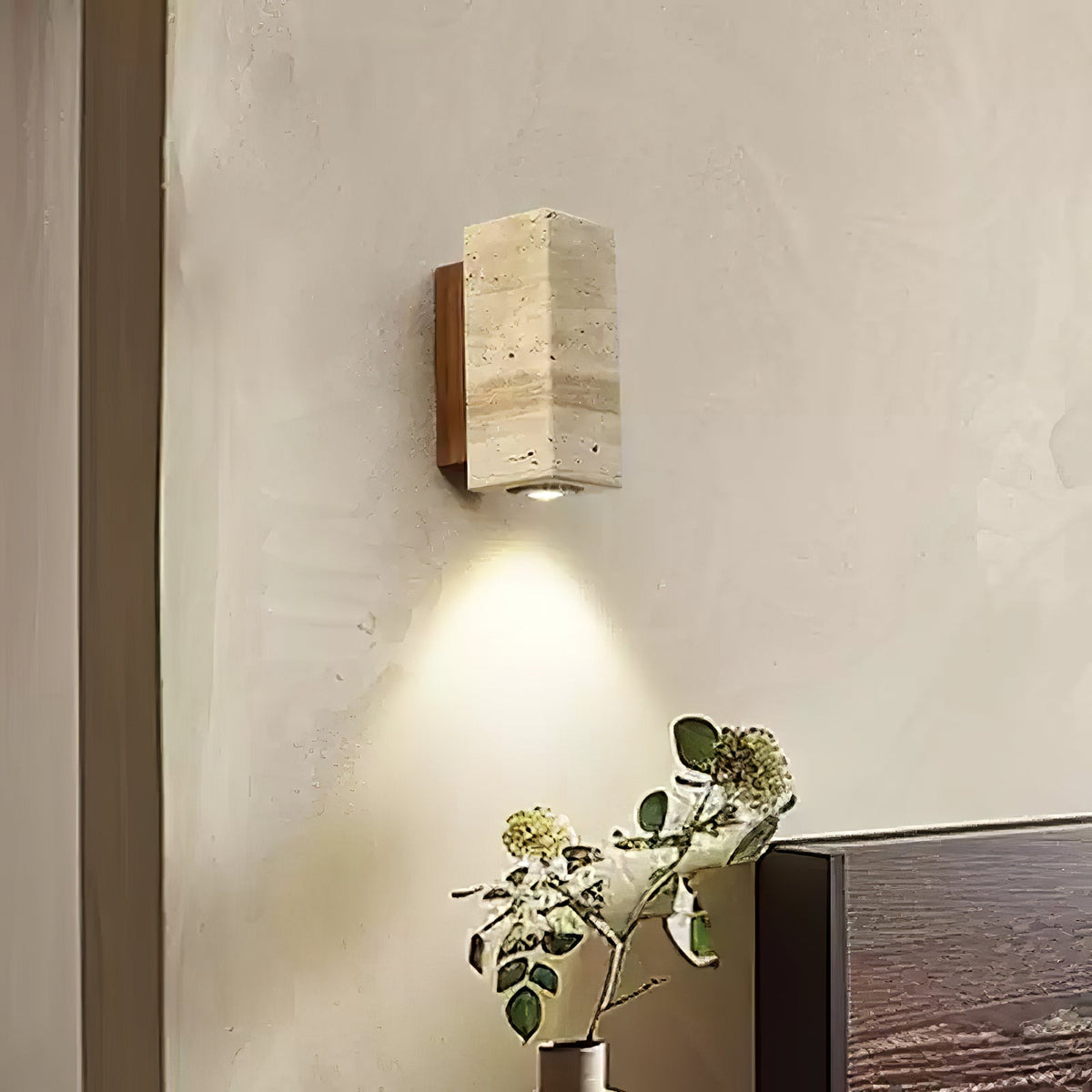 Clérachic travertine wall light lit above a small vase of leaves, mounted on a rough plaster wall beside a dark headboard.